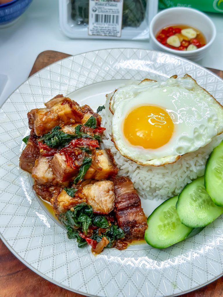 A white plate with a fried egg on top. The plate also contains rice and pork belly. This dish is Pad Kra Pao Moo Grob with Prik Nam Pla, a Thai dish made with stir-fried pork belly, holy basil, and a spicy fish sauce condiment.